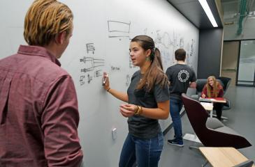 Young woman at whiteboard in classroom, talking to a fellow student